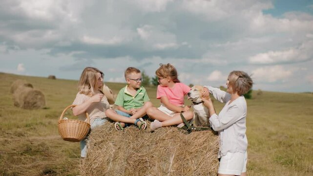 Family enjoys countryside as grandmother feeds dog, child pets its head, woman with basket smiles, children sit on haystack under warm sun, rolling hills and hay bales