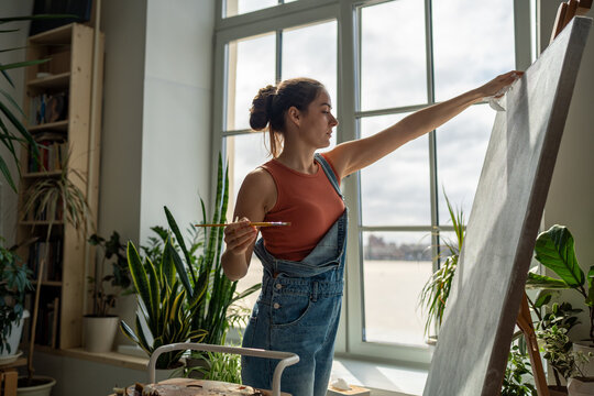 Thoughtful woman artist working on painting. Girl outstretched hand with brush reaches for canvas, making careful thoughtful strokes, creating masterpieces. Working process in art gallery, hobby. 