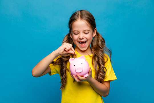 Banking, financial, investment concept - happy laughing little girl puts coins into pink piggy bank on blue background. Pocket money, family budget, savings concept. High quality photo