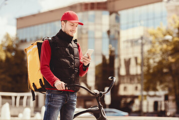 Courier With Phone Using Meal Delivery Application Standing With Bicycle Outside In Urban Area....