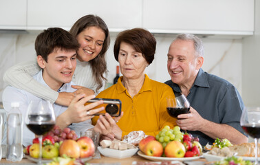 Joyful young guy and girl enjoying family dinner, sharing digital memories from smartphone with smiling senior parents in cozy kitchen. Generation bonding concept