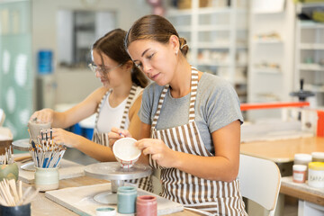 Young female visitors of master class for manufacture of ceramic memorabilia perform work on molding blanks from raw clay and painting staining them from afar before firing them.