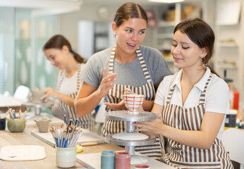 Young female teacher explains to girl student how to paint ceramic cup in workshop..