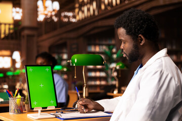 Future healthcare professional studying medical methods next to chroma key screen in an educational library, reading case files and academic papers. Black man works on academic growth.
