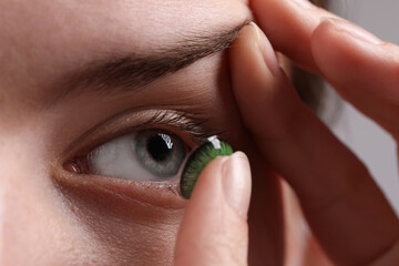 Woman putting in green color contact lens on grey background, closeup