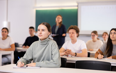 Group of school teenagers with pens and notebooks studying in classroom with teacher
