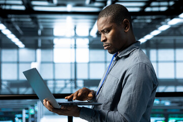 African american engineer in data center with laptop automating routine maintenance tasks,...