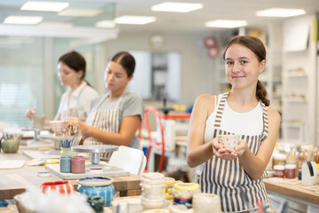 Teenage girl student in apron with handmade ceramic cup posing in ceramics workshop