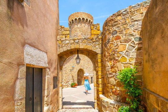 A young blonde female tourist walks through the interior archways and gates of the 12th century Murallas de Tossa de Mar fortress in the Costa Brava town of Tossa de Mar, Spain.