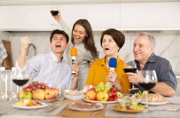 Joyful elderly man and woman sharing microphones with adult children and singing passionately during festive home gathering with food and drinks