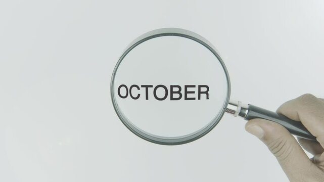 October text or word highlighted by a magnifying glass in a hand on a white background, symbolizing focus on the month, seasonal changes, and events related to autumn