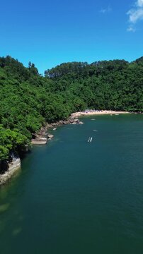 Aerial view of Praia do Cedro located in Ubatuba, Brazil. People enjoy the beach and the sea water crashes against the rocks on the coast. Green trees are seen in the beautiful landscape.