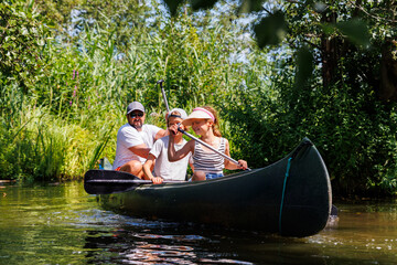 Man with two children enjoy having fun paddling canoe rent across lake river water wearing bright orange life vests hot sunny summer day. Healthy recreational activity lifestyle vacation trip journey