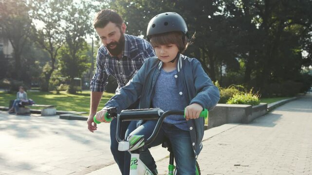 Pleasant-looking young Caucasian dad with beard helping kid to ride a bicycle, holding bike. Little boy riding through the park. Summertime. Outdoors