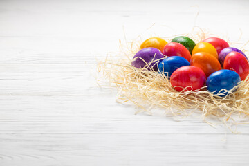 Multicolored Easter eggs lie in a pile in hay on a white wooden table