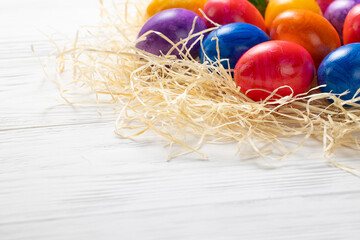 Multicolored Easter eggs lie in a pile in hay on a white wooden table