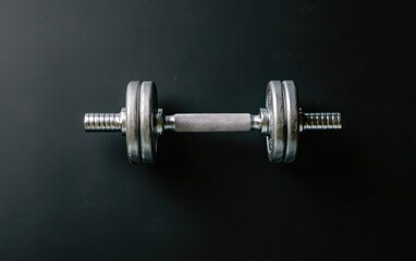 Overhead shot of a single chrome dumbbell on a dark gray textured surface. The dumbbell is the main focus, set against a minimalist background
