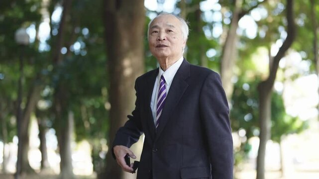 A 78-year-old Japanese man in a suit stands and operates a smartphone in a natural park filled with trees