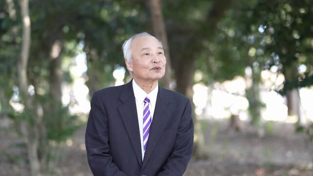 A winter park in Japan. A 78-year-old Japanese man in a suit stands and talks on top of the fallen leaves in a natural park filled with trees.