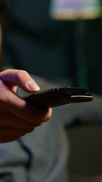 Vertical video Close up of device used by person to scroll through streaming content, picking movie. Focus on remote control used by man in blurry background switching channels on television screen