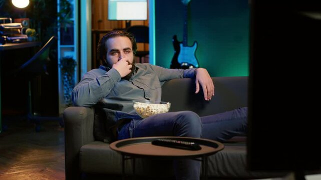 Homeowner bored at home munching on snacks, using TV to binge series on subscription based streaming services. Man sitting on couch, wasting time with VOD shows on TV, eating popcorn, camera A