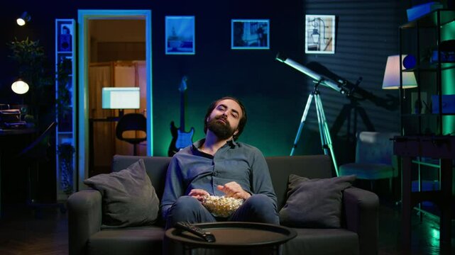 Fatigued man watching shows on TV late at night and munching on popcorn, paying close attention to screen. Tired sedentary man sitting in pajamas all day consuming mindless entertainment, camera B