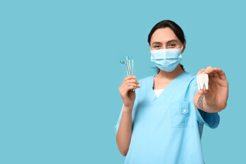Young female dentist in mask with dental tools and tooth model on blue background