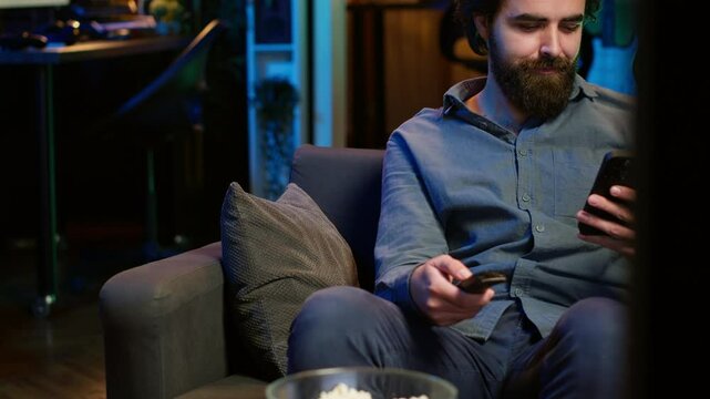 Smiling person on couch picking movie on streaming service using remote and texting on mobile phone. Happy man watching shows on television, zapping though channels, chatting smartphone, camera A