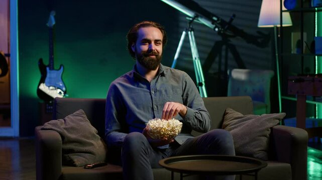 Man turning on TV using remote control, enjoying leisure time at home, munching on popcorn. Couch potato person in neon illuminated apartment eating salty snack, entertained by television, camera A