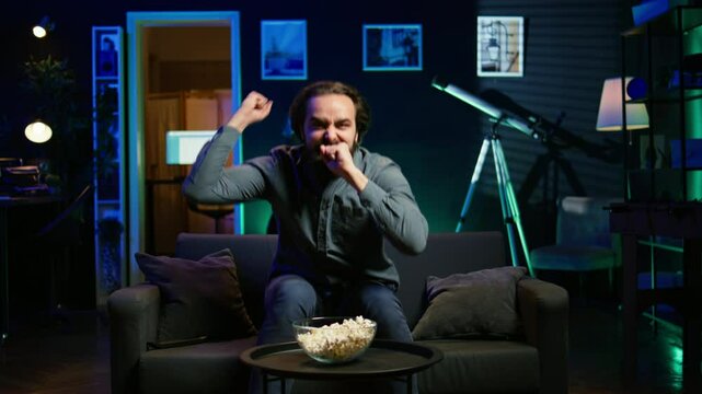 Enraged supporter watching sports match on TV, flabbergasted by goal conceded by favorite team. Appalled man looking at football competition, reacting in anger to player missing penalty, camera B