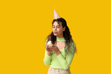 Beautiful young African-American woman in party hat holding birthday cupcake with burning candle on yellow background