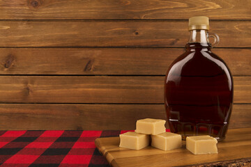 Bottle of rich maple syrup with golden fudge pieces on a wooden surface, set against a rustic wooden backdrop. A warm, inviting scene capturing the essence of Canadian cuisine and sweet indulgence.