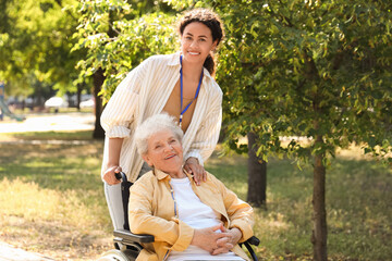 Young African-American female medical worker walking with elderly woman on wheelchair outdoors