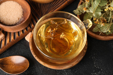 Cup of tasty linden tea with sugar and spoon on black background