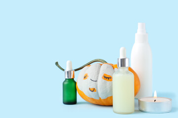 Pumpkin with drawn face, clay mask, candle and bottles of cosmetic products on blue background