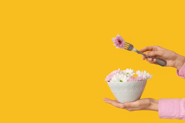 Female hand holding bowl and fork with beautiful flowers on yellow background