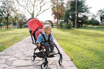 Little girl sits in a stroller on a path and peers among the trees in a green park