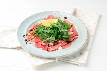 Delicious beef carpaccio with arugula, cheese, balsamic vinegar and lemon on white wooden table, closeup