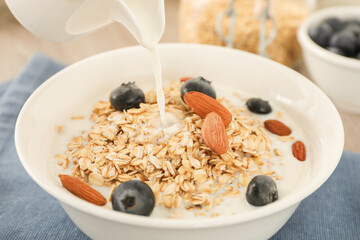 Pouring milk into bowl with oatmeal, blueberries and almonds at table, closeup