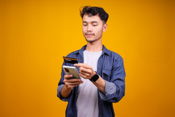 Fototapeta premium Smiling Asian Man Holding Credit Card And Mobile Phone While Standing Against Yellow Background