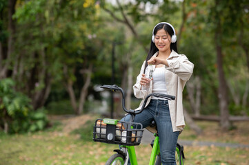 A smiling asian woman is opening a clear water bottle while riding a green bicycle with headphones...