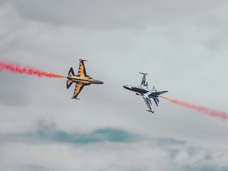 wo supersonic jets perform a breathtaking high-speed crossover at the Avalon Airshow, leaving vibrant red smoke trails against a cloudy sky. Precision, skill, and adrenaline in an electrifying moment.