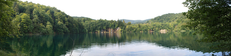Beautiful wide landscape - blue lake in evergreen forest under fluffy clouds in the sky on a sunny day, with green grass in the foreground