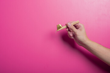 Asian hand holding a traditional canting tool used in batik making, with a wooden handle and brass bowl tip, against a pink background. Represents heritage, craft, and Indonesian culture.