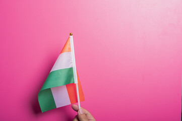 Asian hand holding the Indian national flag with orange, white, and green stripes and the Ashoka Chakra, set against a pink background. Represents patriotism, culture, or celebration of India.