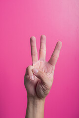 Asian hand showing three fingers raised, thumb and pinky folded, against pink background,...