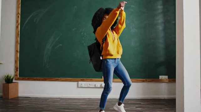 A joyful Black girl, approximately 10 years old, with curly hair, wearing a yellow hoodie and jeans, dances carefree in a bright classroom amid a green chalkboard background.