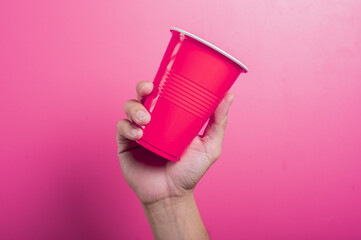 Asian hand holding a red plastic party cup against a vibrant pink background. The hand is...