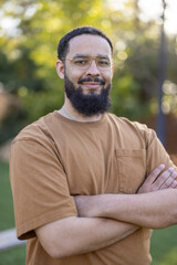A confident man with crossed arms and an inviting smile stands proudly in a vibrant outdoor environment, his relaxed posture and warm demeanor welcoming others to engage and connect with him