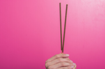 Asian hands holding three brown incense sticks vertically against a pink background. The gesture implies ritual, meditation, spirituality, or traditional cultural practices and offerings.